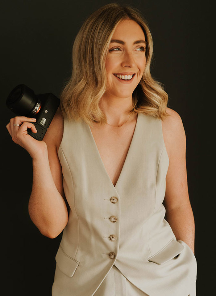 a smiling person wearing a beige waistcoat and trousers holding a camera and looking to the side