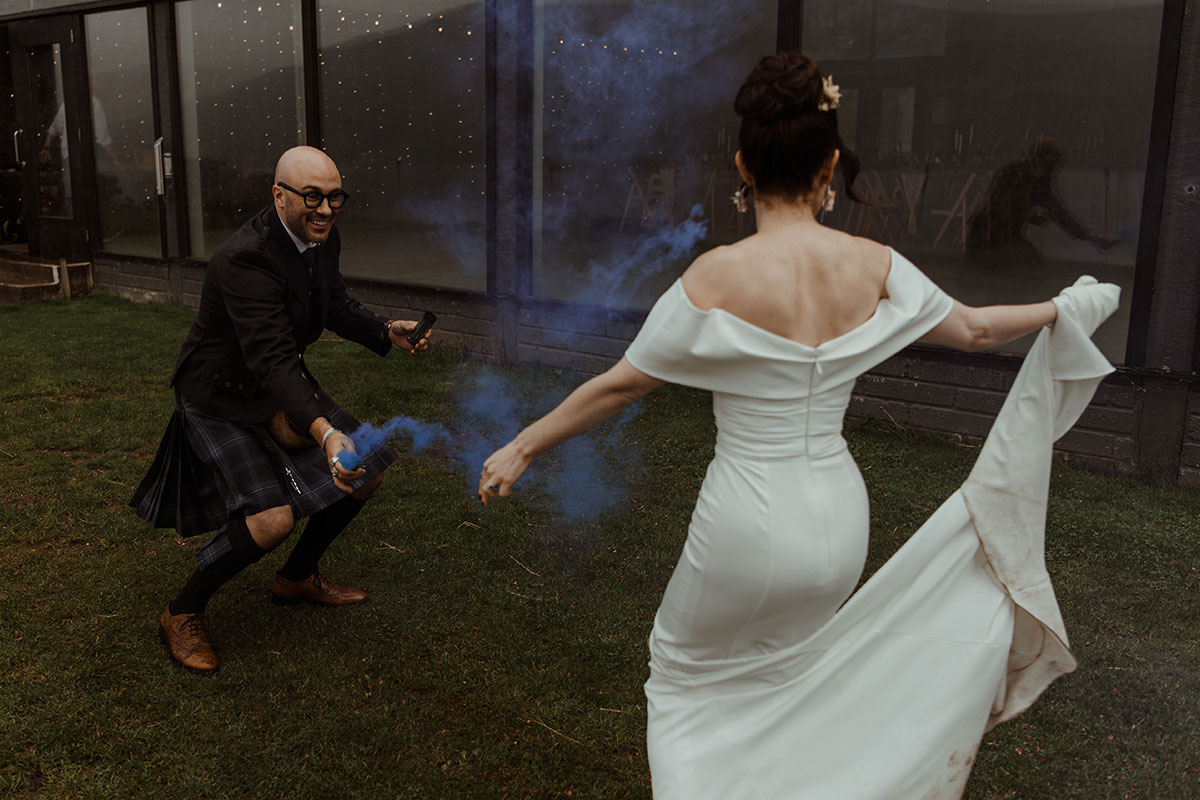 groom in blue, grey and white tartan kilt outfit crouches down on grass holding blue smoke stick as bride in white wedding dress dances in front