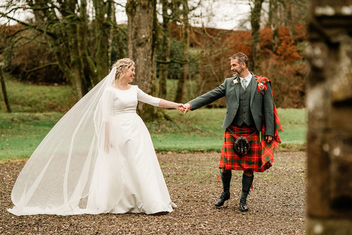 bride in white wedding dress and long veil and groom in red and green tartan kilt stand outdoors hand in hand, facing each other with that one arm outstretched between them