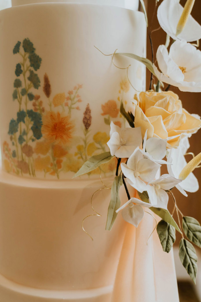 A close up of a wedding cake with a flower design on the icing as well as real flowers on the side of the cake