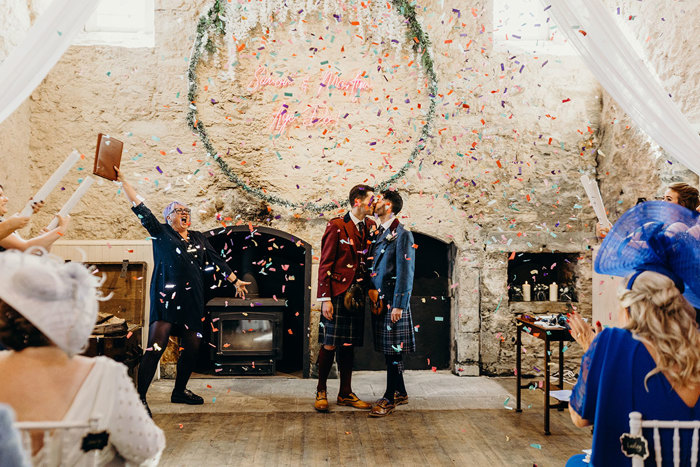 two grooms in kilts kissing while being showered in colourful confetti in a room with rustic white wall. A celebrant cheers on the left.