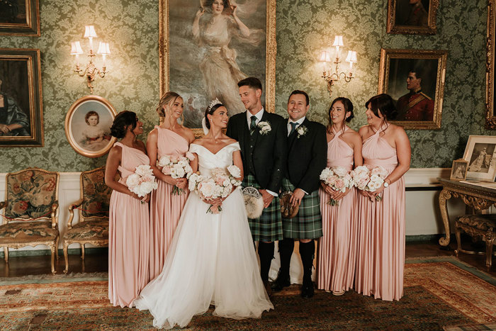 A wedding party stands together in a room with green wallpaper and paintings on the wall 