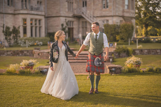 A bride wearing a white dress and a leather jacket holds hands with a groom wearing a red kilt walking across a lawn