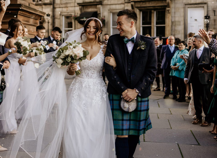 Bride and groom walk outside their wedding venue as loved ones throw flower petals at them