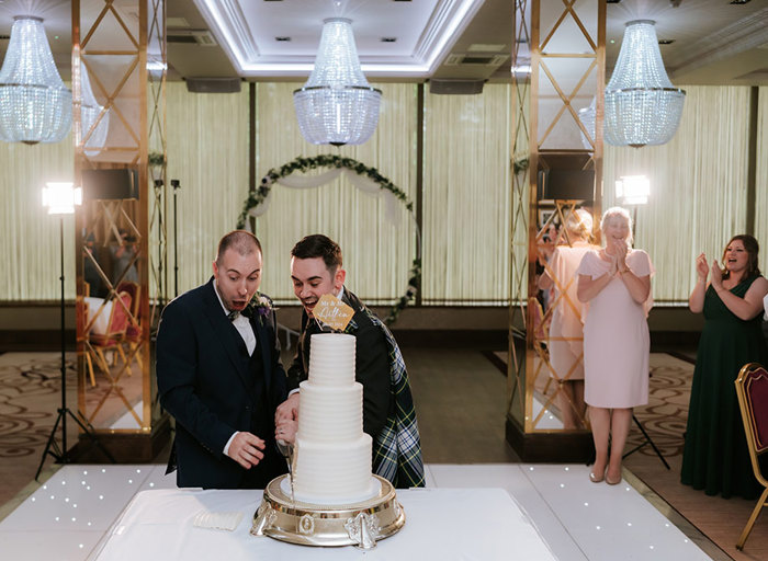 two grooms looking surprised as they cut a three-tier wedding cake. There is a white light-up dance floor and some guests are clapping in the background