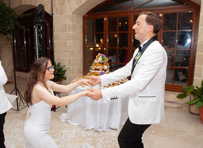 a man wearing a smart white jacket dancing with a young girl wearing a white dress. There is a wooden glazed door and a table with a cannoli tower on it in background