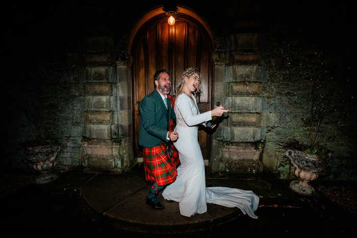 groom in red and green tartan kilt outfit stands directly behind bride in white wedding dress and veil as they spray a bottle of champagne to the side