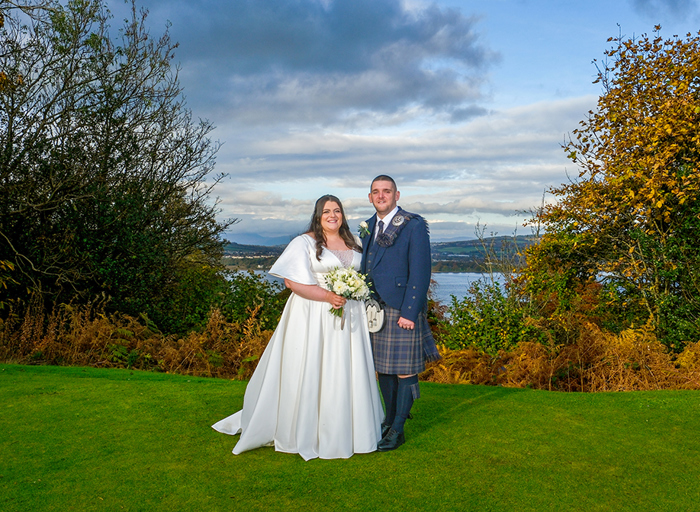 A bride and groom stand next to each other facing the camera with a view of hills and water behind them