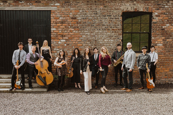 a line up of thirteen musicians holding a variety of instruments, all smiling and stood outside in front of a red brick wall