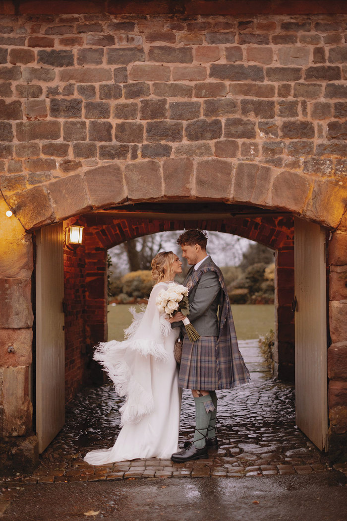 blonde bride in feathery cape and white wedding dress holds bouquet of white roses and stands smiling and facing groom in green jacket and red, blue, green kilt with matching fly plaid, both stood under outdoor stone archway