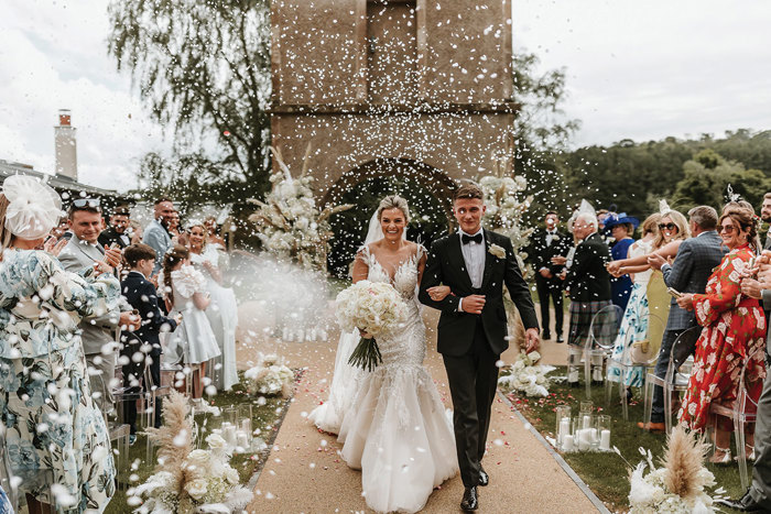 bride and groom walk up outdoor aisle surrounded by guests in rows of clear chairs as white confetti falls around them 