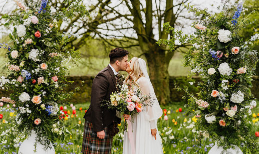 a bride and groom kissing in a garden filled with abundant colourful flowers. Two large white urns filled with tall pastel and foliage flower arrangements flank them 