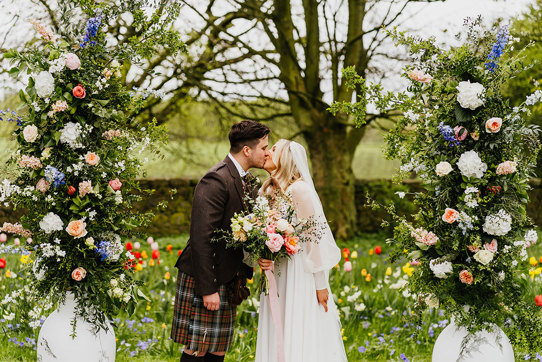 a bride and groom kissing in a garden filled with abundant colourful flowers. Two large white urns filled with tall pastel and foliage flower arrangements flank them 