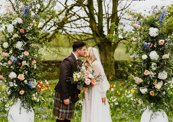 a bride and groom kissing in a garden filled with abundant colourful flowers. Two large white urns filled with tall pastel and foliage flower arrangements flank them 