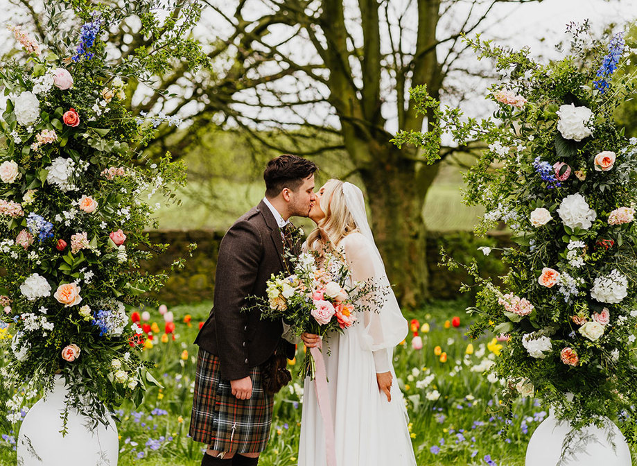 a bride and groom kissing in a garden filled with abundant colourful flowers. Two large white urns filled with tall pastel and foliage flower arrangements flank them 