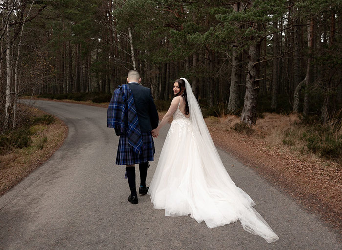 a bride and groom walking hand in hand on a path in a forest setting.