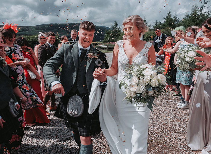 a bride and groom walk through two rows of guests throwing confetti at them in a countryside setting