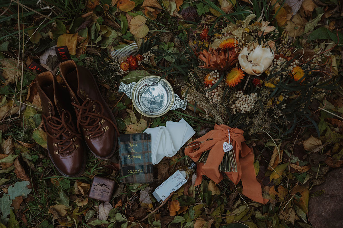 a flat lay of wedding details on leaves including silver engraved quaich, tartan ribbon, wedding rings in box and bouquet of rustic style flowers