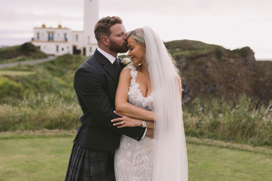 Groom kisses brides forehead as they pose in outdoor photoshoot