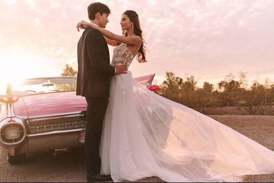 Model wearing corset bodice flowy skirt wedding dress in front of pink corvette with groom next to her