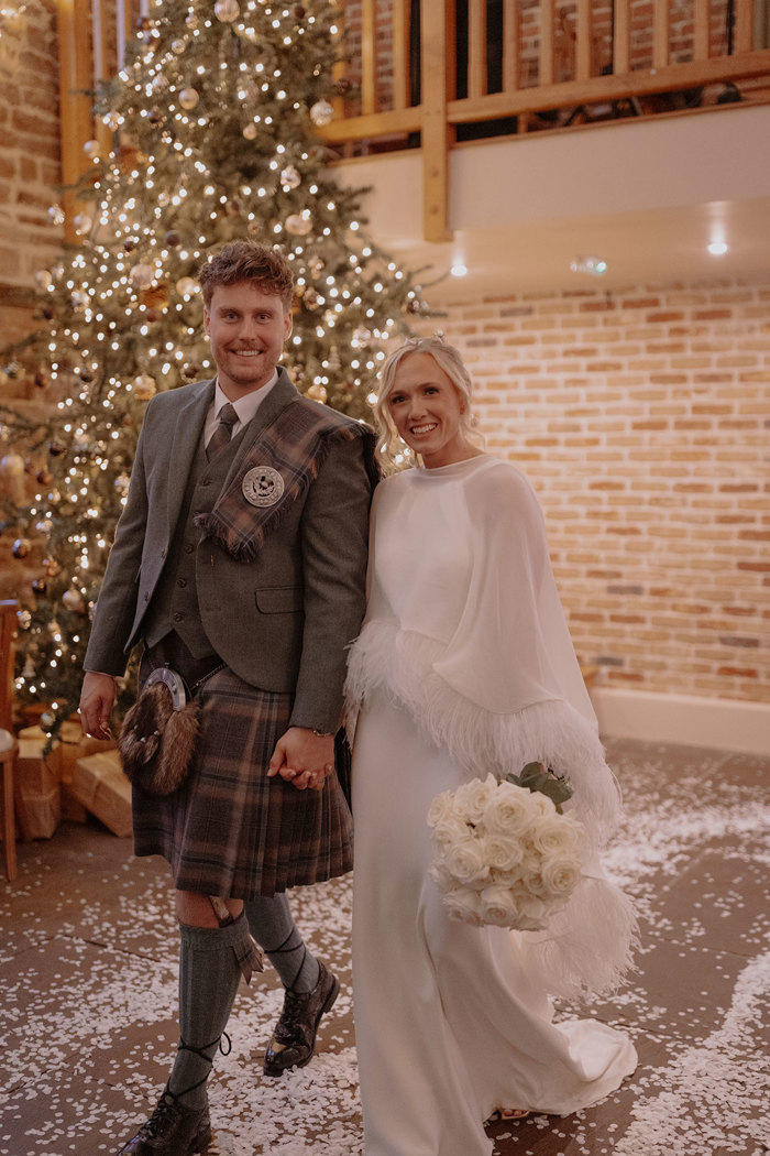 blonde bride in feathery cape and white wedding dress holds bouquet of white roses down by one side and holds groom's hand in the other as he wears green jacket and red, blue, green kilt with matching fly plaid, both walking by Christmas tree and a confetti-covered floor
