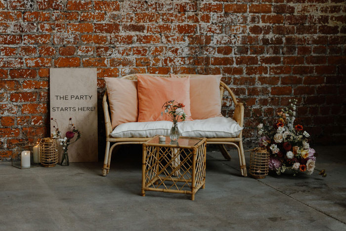 a wicker love seat with coral and pastel pink cushions is surrounded by small wildflower floral arrangements and a sign that says 'The party starts here'