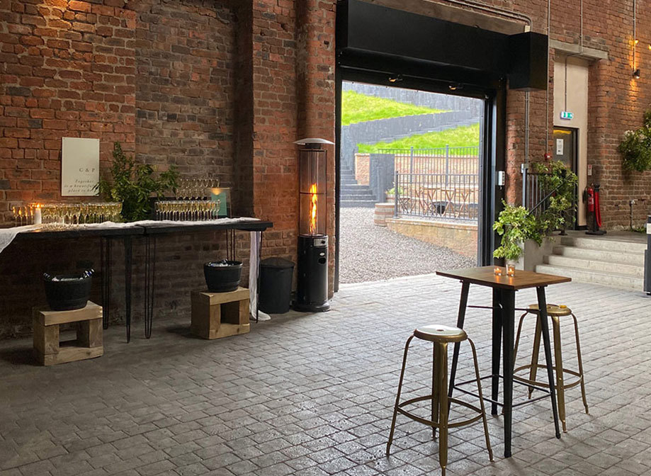 A room with brickwork walls and floors and a large roller door with a small tall table and stools closer to the camera