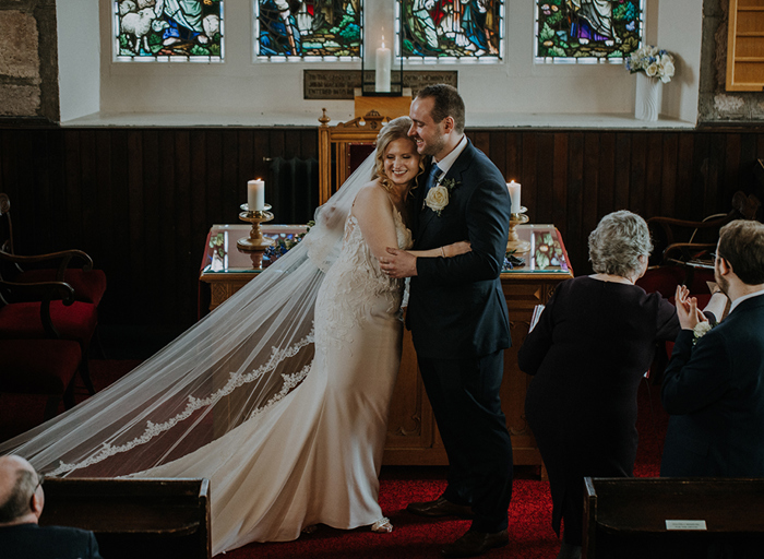 A bride in a fitted dress and long veil and groom in a suit hugging at the alter 