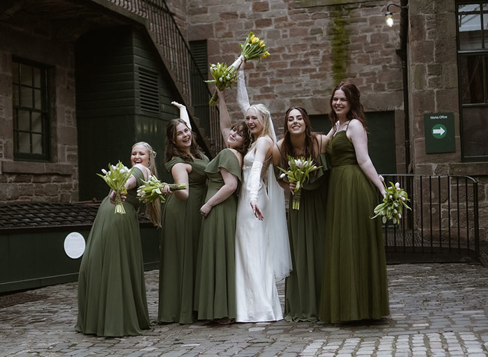 a bride holding a bunch of yellow tulips in the air with five bridesmaids wearing dark green dresses posing. They are standing on a cobbled pavement and there is a brown brick building and staircase in the background