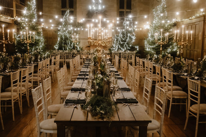 long tables set for dinner with illuminated Christmas trees in the background and festoon lighting hanging above them.