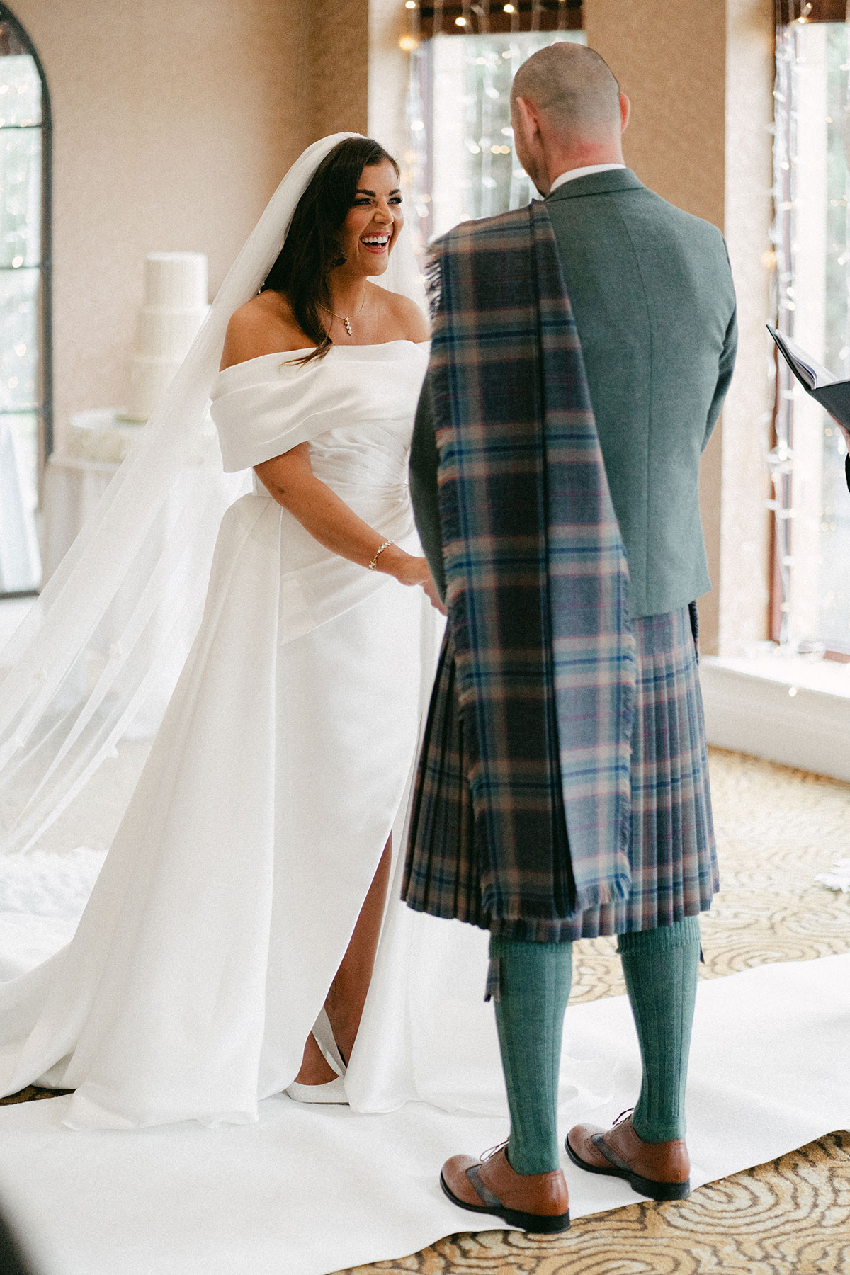 bride with long veil and off the shoulder wedding dress with a leg slit stands across from a groom in green, blue and purple tartan kilt outfit with green jacket holding his hands 