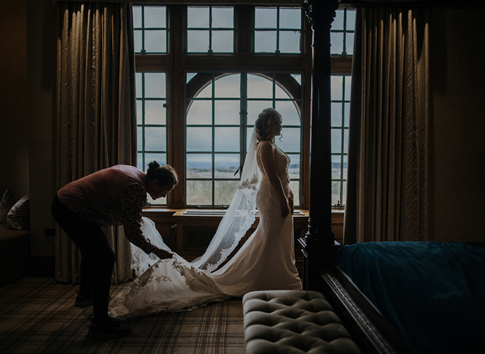 a woman adjusts a bride's long veil as they are both backlit by the light coming in from the large window behind them