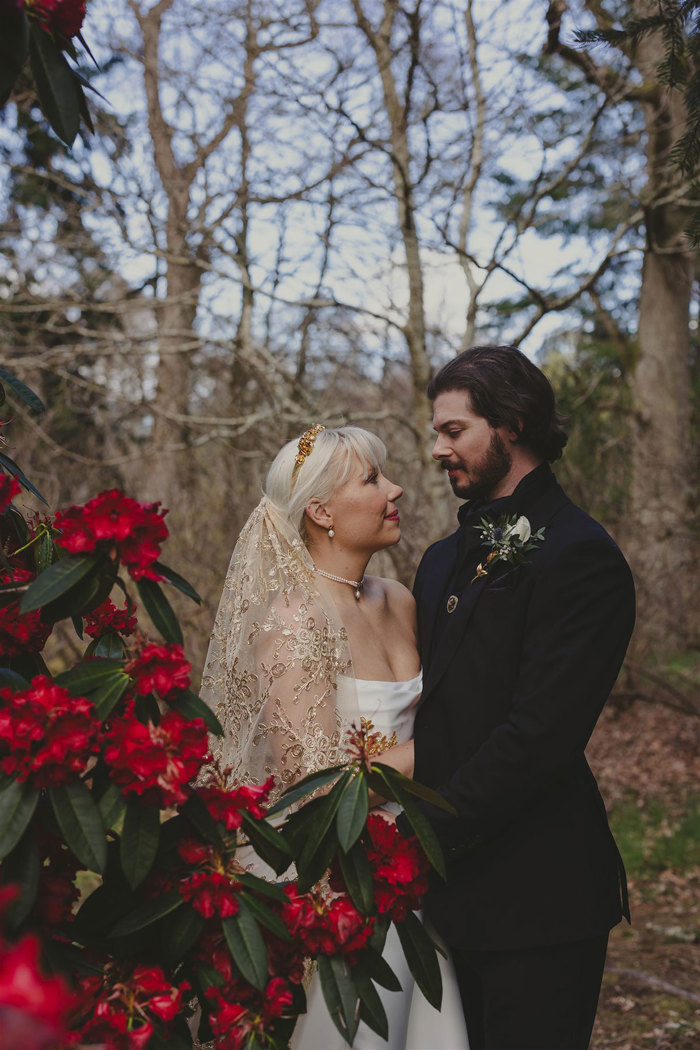 a bride and groom posing with trees in the background and red flowers in the foreground.