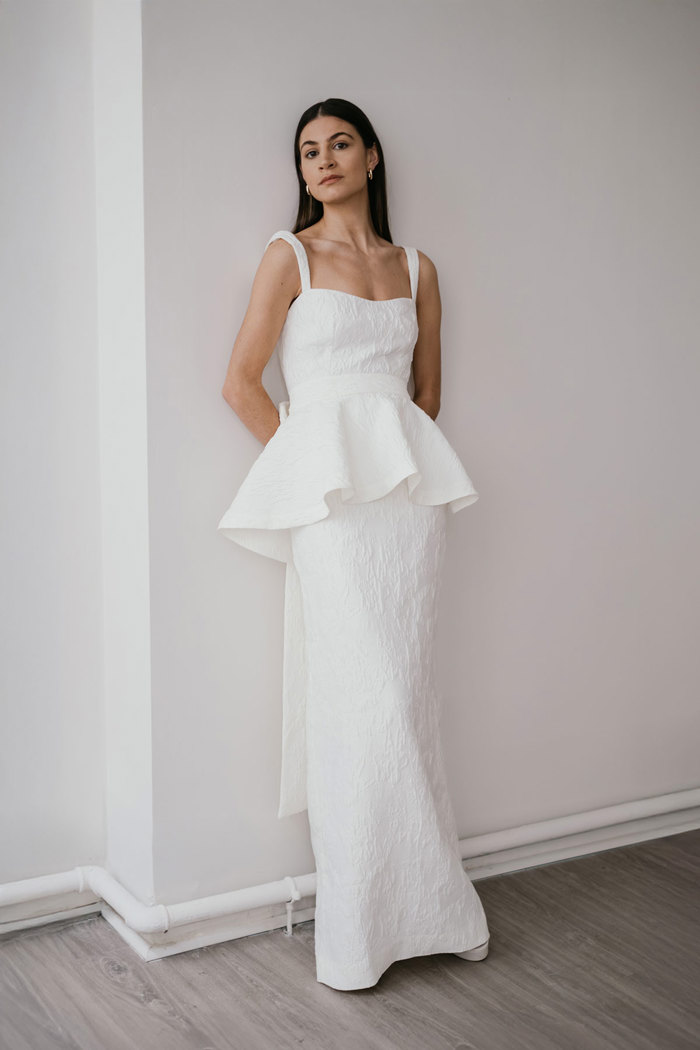 dark haired model leans her back against plain white wall, hands tucked behind her back as she wears a white textured wedding dress with a peplum waist