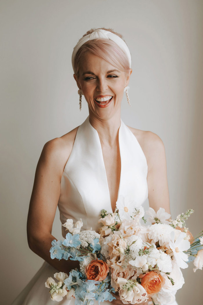 short pale pink haired bride wears white headband and v-neck wedding dress while holding a pastel blue, pink and white bouquet