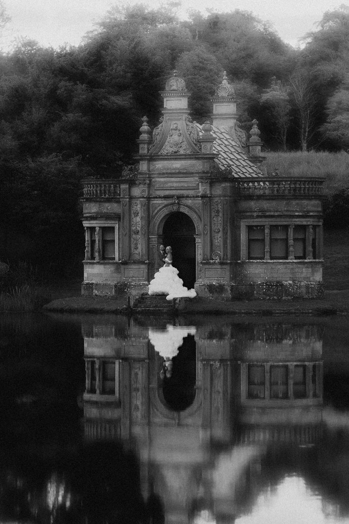 A black and white photo of a bride and groom standing in the doorway of a small building across a body of water