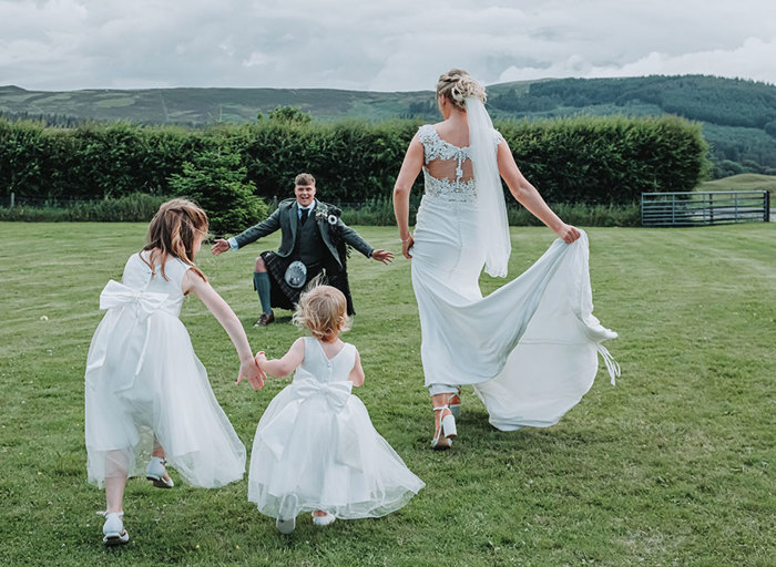 two flower girls and a bride running over grass towards a man in a kilt with outstretched arms