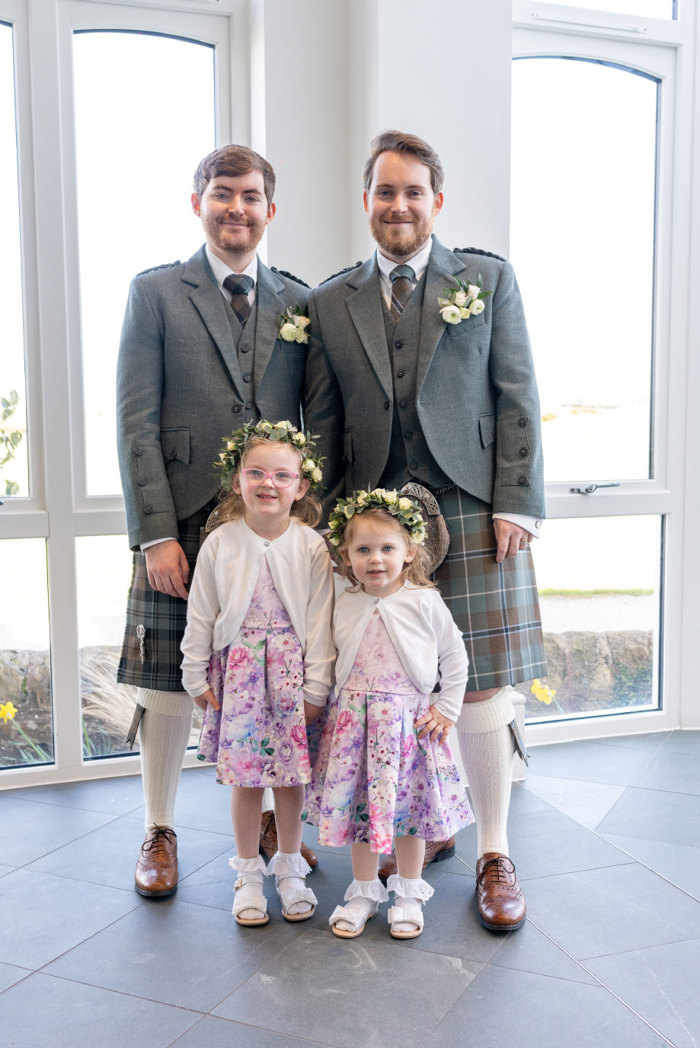 two grooms wearing kilts posing with two girls wearing pink flower dresses and white cardigan in front of windows