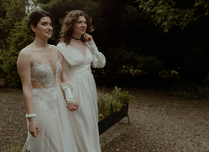 two brides walking hand-in-hand on a gravel path in garden filled with dark green bushes and trees