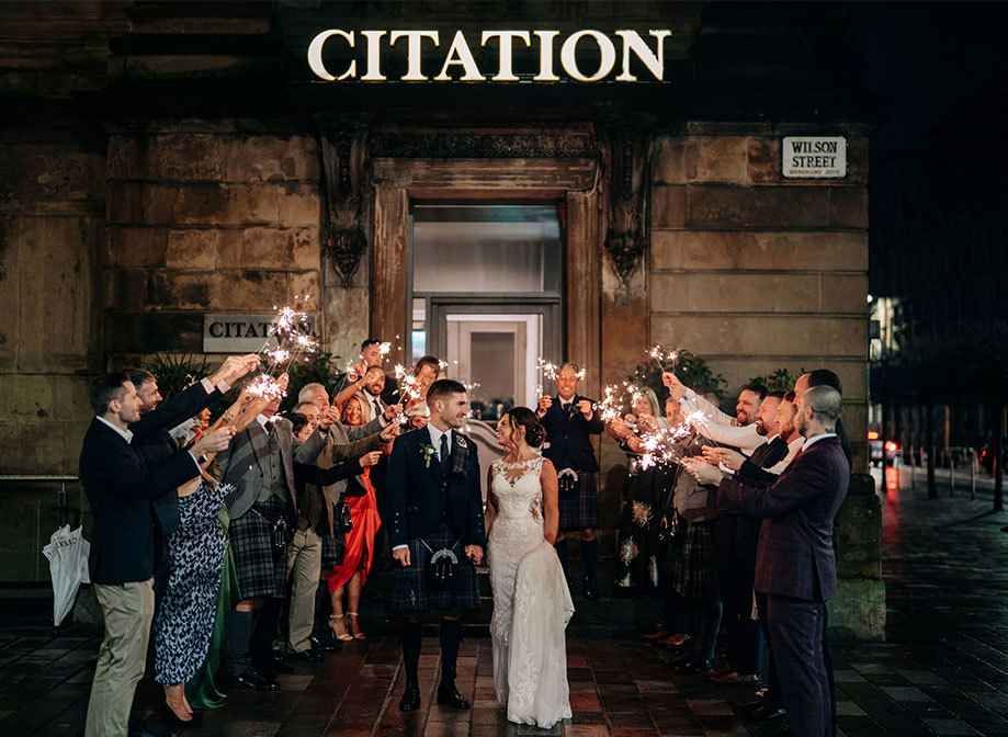 A bride and groom looking at each other as they walk through a crowd of people holding sparklers