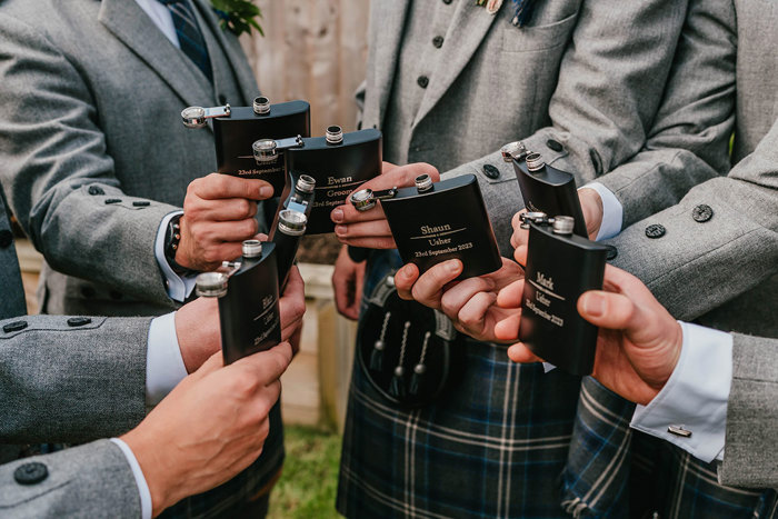 Seven men in grey kilts hold personalised black hipflasks close together 