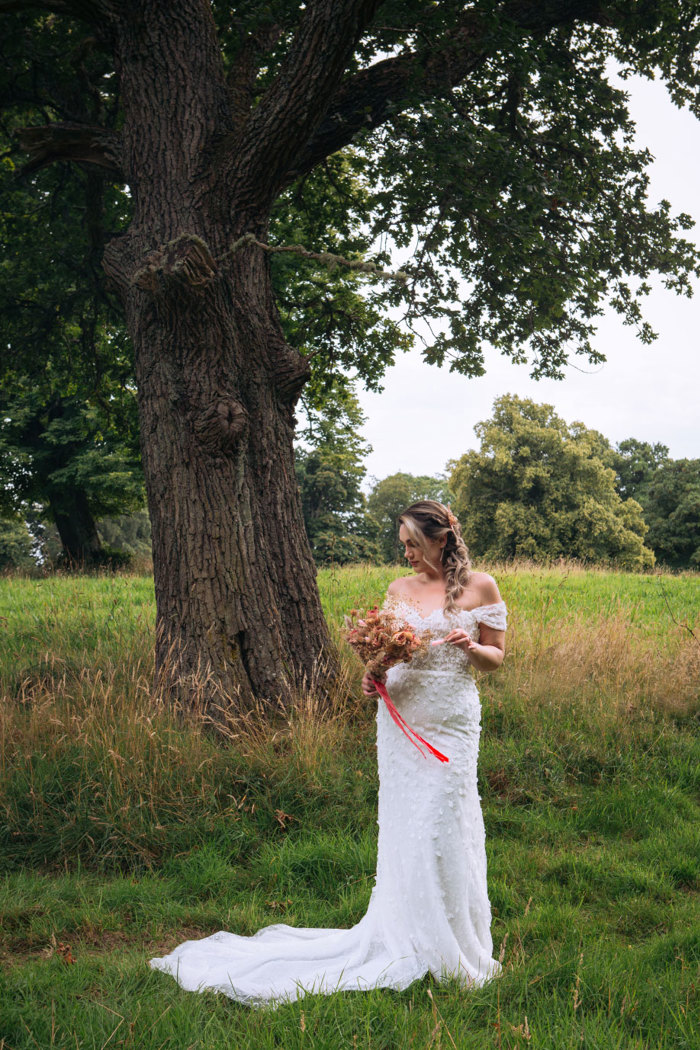 bride stands in a grassy field wearing an off the shoulder strapless wedding dress and holding a dried floral bouquet