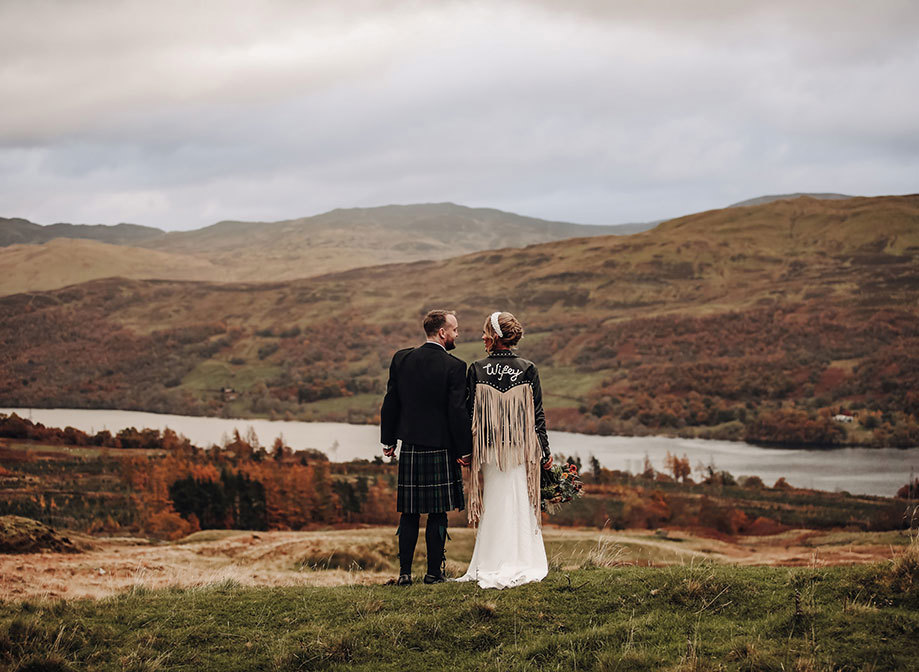 a bride wearing black fringed leather jacket holds hands with a groom wearing a kilt as they look at each other amid autumn moorland in Scotland with a loch and hills in background