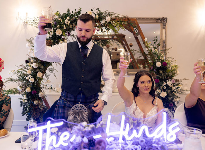 a standing groom raises a glass to toast next to seated bride. There is a neon sign on table reading 'The Hynds' and a wooden hexagonal frame covered with flowers behind them