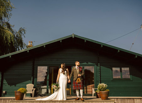 A bride and groom stand in front of a small green building holding hands 