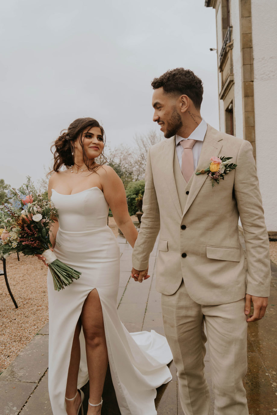 A bride in a long dress with a slit holding a colourful bouquet holds hands with a groom in a beige suit 