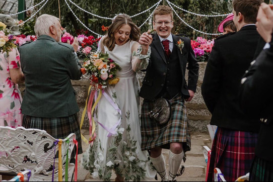 A bride and groom smiling as they walk down the aisle holding hands with their guests on either side clapping 