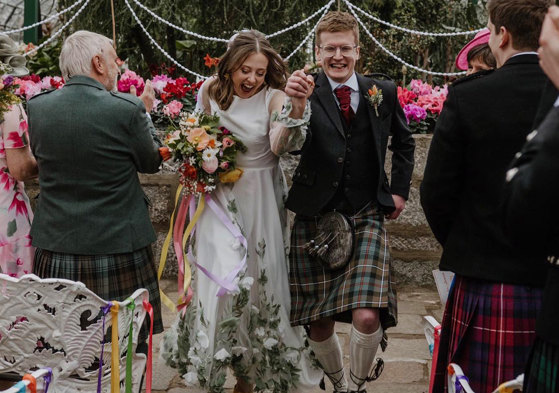 A bride and groom smiling as they walk down the aisle holding hands with their guests on either side clapping 