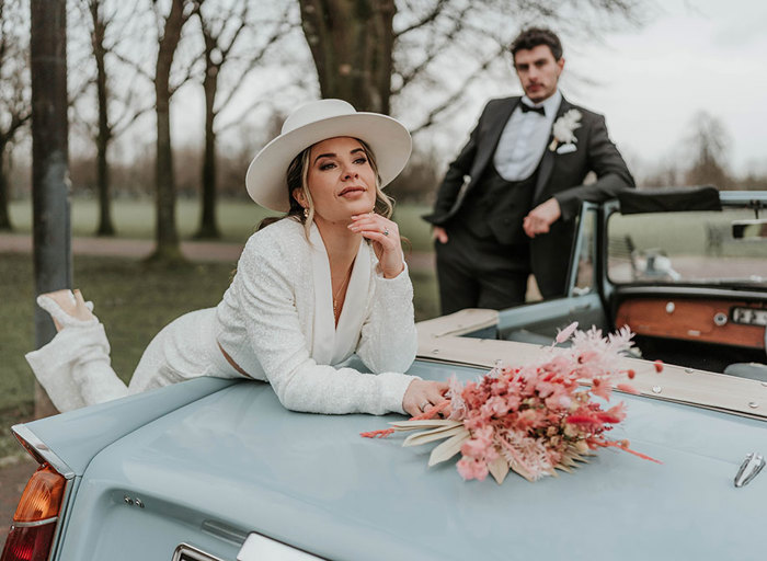 A woman wearing a white suit and white hat leans over the book of a blue convertible car and a man in a suit leans on the windshield in the background