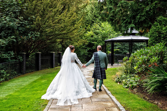 a bride and groom walking hand-in-hand through a garden towards a black Victorian bandstand.